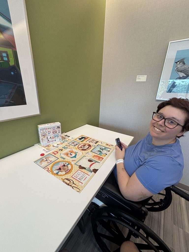 A young woman sits at a table in a wheelchair smiling as she completed a puzzle with different dogs on it.